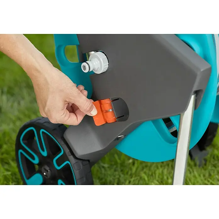 Gardena Schlauchwagen CleverRoll M Easy - Kunststoff/Metall, Schlauchkapazität: 60 m, 13 mm (½″) - Nahaufnahme einer Hand, die einen orangefarbenen Verschluss an einer Gardena-Schlauchtrommel betätigt.