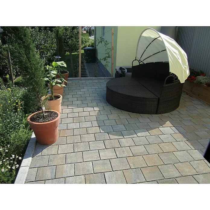 Diephaus Pflasterstein Caracas - Muschelkalk, 21 x 14 x 8 cm, Rechteckig - Ein einladender Außenbereich mit einem runden Rattan-Sofa mit Sonnendach und Pflanzen in Terrakotta-Töpfen auf einem gepflasterten Boden.