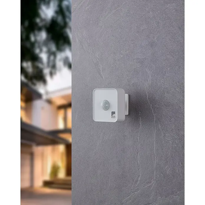 Eglo Connect LED-Lampe - Ausstrahlungswinkel: 120 °, IP44, 7,5 x 7 x 7,5 cm - Nahaufnahme eines weißen EGLO-Bewegungsmelders an einer grauen Wand, mit einem verschwommenen Gebäude im Hintergrund.