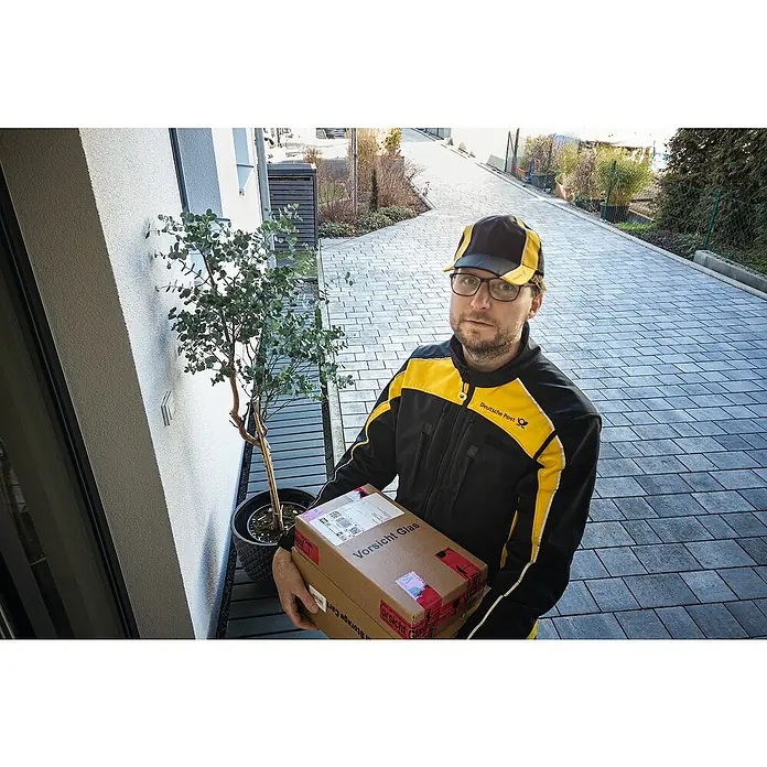 Abus Überwachungskamera PPIC46520 BK - IP66 - Ein Postbote der Deutschen Post liefert ein Paket "Vorsicht Glas" vor einer Haustür aus. Zu sehen sind die Aufschrift und das Logo der Post.