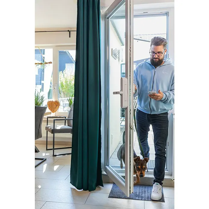 Abus Funk-Türschlossantrieb Wintecto one FCA4100 W - Geeignet für: Fenster, Weiß, Bedienung: Über App - Ein Mann verlässt mit seinem Hund das Haus, wobei ein smartes Türschloss am geöffneten Fenster sichtbar ist. Er benutzt sein Smartphone.