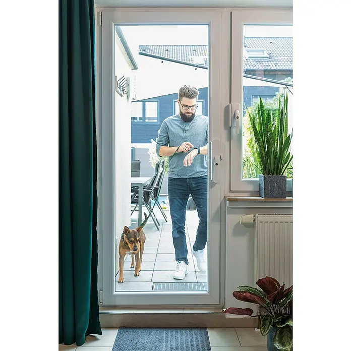 Abus Funk-Türschlossantrieb Wintecto one FCA4100 W - Geeignet für: Fenster, Weiß, Bedienung: Über App - Ein Mann mit Hund steht in einer Tür und schaut auf seine Smartwatch. Helle, moderne Tür mit großzügiger Glasfläche.