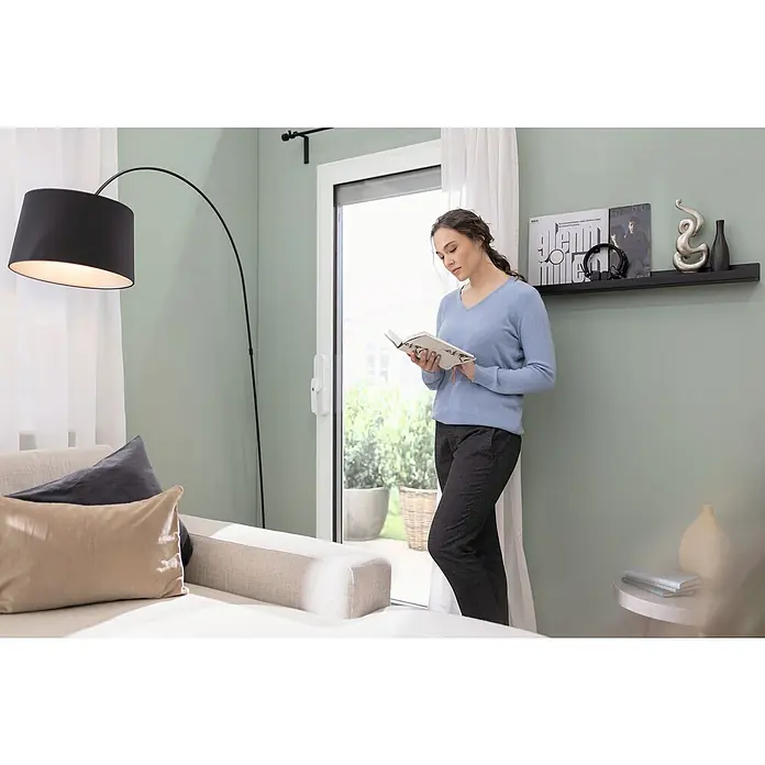 Abus Funk-Türschlossantrieb Wintecto one FCA4100 W - Geeignet für: Fenster, Weiß, Bedienung: Über App - Eine Frau liest in einem hellen Wohnzimmer ein Buch, neben einer modernen Stehlampe und einem Sofa mit Kissen.