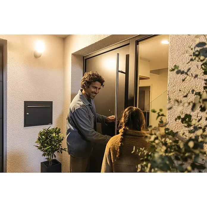 Abus Überwachungskamera PPIC46520 BK - IP66 - Einladende Haustürszene: Ein Mann öffnet eine moderne Tür mit langem Griff, während eine Frau eintritt. Außenwand mit Briefkasten und Beleuchtung.