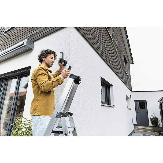 Abus Überwachungskamera PPIC46520 BK - IP66 - Ein Mann auf einer Leiter befestigt eine intelligente Außenleuchte an einer Hauswand und bedient sie mit seinem Smartphone.