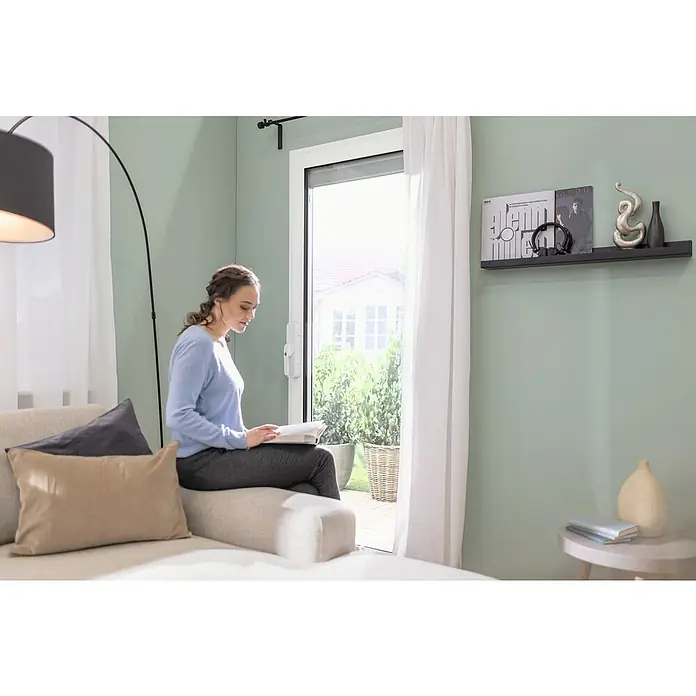 Abus Funk-Türschlossantrieb Wintecto one FCA4100 W - Geeignet für: Fenster, Weiß, Bedienung: Über App - Eine Frau sitzt auf einem Sofa und liest ein Buch. Im Hintergrund ist eine Tür mit Blick auf einen Garten zu sehen.