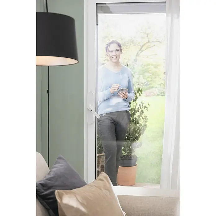 Abus Funk-Türschlossantrieb Wintecto one FCA4100 W - Geeignet für: Fenster, Weiß, Bedienung: Über App - Das Bild zeigt eine moderne Tür mit einem eleganten Türgriff, während eine Frau im Hintergrund ihr Smartphone benutzt.