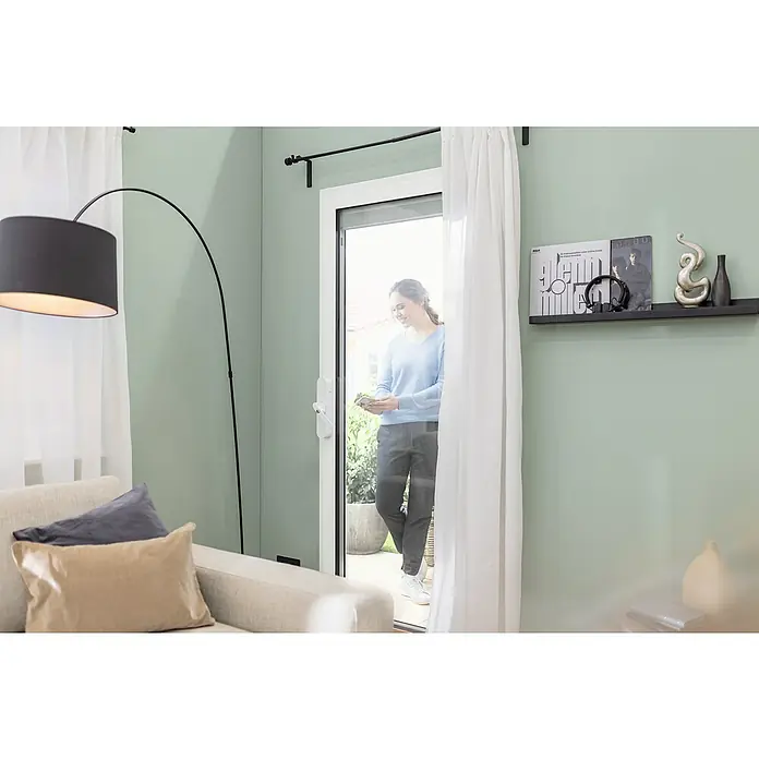 Abus Funk-Türschlossantrieb Wintecto one FCA4100 W - Geeignet für: Fenster, Weiß, Bedienung: Über App - Eine Frau steht an einer Terrassentür mit Insektenschutzgitter. Es ist mit einem weißen Rahmen und Griff ausgestattet.