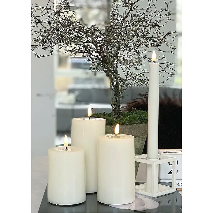 DeluxeHomeart LED-Kerze Indoor - Weiß, Ø x H: 7,5 x 15 cm, Echtwachs - Kerzenensemble: Drei Stumpenkerzen und eine Stabkerze in einem modernen, würfelförmigen Kerzenhalter, umgeben von einem dekorativen Zweig.