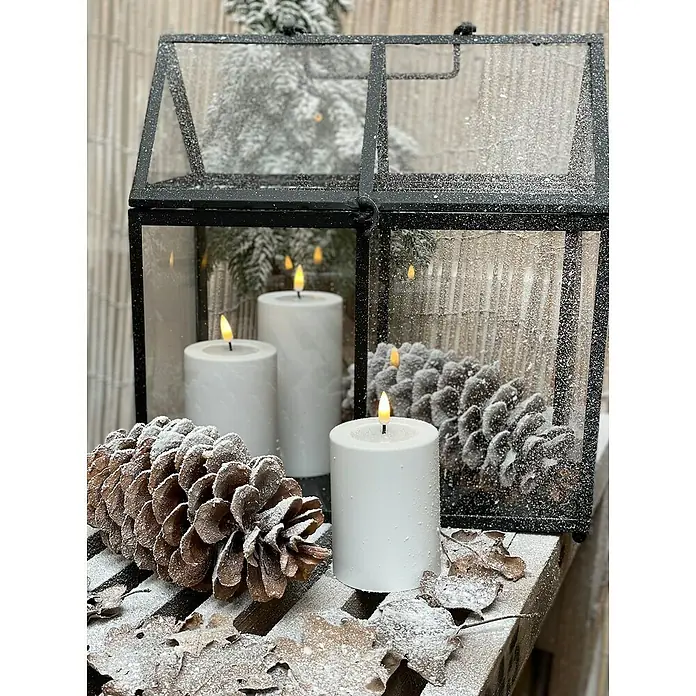 DeluxeHomeart LED-Kerze Indoor - Weiß, Ø x H: 7,5 x 15 cm, Echtwachs - Stimmungsvolles Arrangement mit weißen Kerzen und Tannenzapfen, dekoriert mit Kunstschnee, in einer schwarzen Laterne für weihnachtliche Atmosphäre.