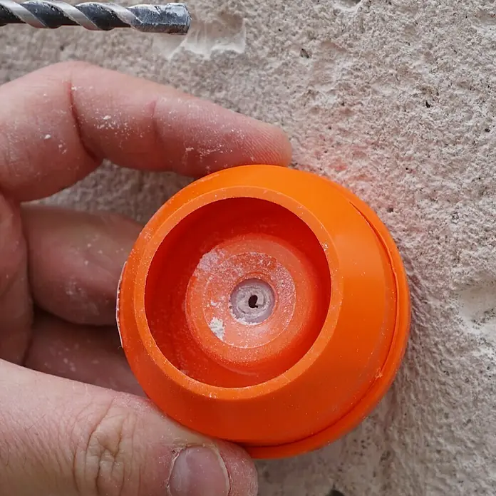 Wolfcraft Bohrstaubfänger - Durchmesser Bohrer: 4 mm - 8 mm, Kunststoff - Nahaufnahme einer orangefarbenen Bohrschablone, die an einer Wand befestigt wird, um präzise Löcher zu bohren. Staub umgibt die Vorrichtung.
