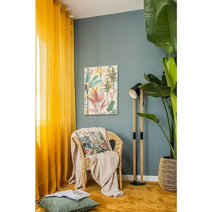 Cojín Pana - 45 x 45 cm, Poliéster, Verde, Cuadrado - Un interior acogedor con una silla de ratán, arte de plantas en la pared, una lámpara de pie y una gran planta de interior.
