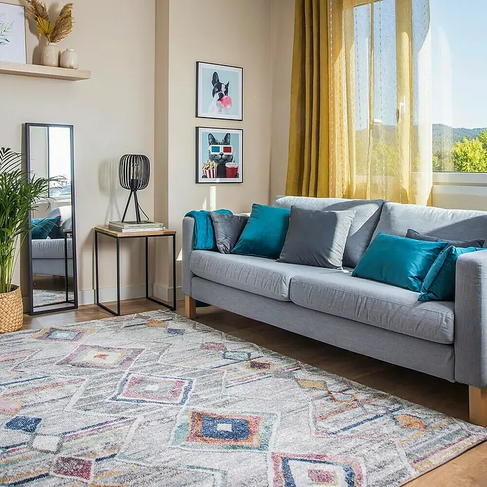 Cojín Velvet - 50 x 30 cm, Textil, Turquesa, Rectangular - Un sofá gris claro con cojines azules y grises, una alfombra estampada y decoración. El sofá ofrece comodidad en un espacio habitable moderno.