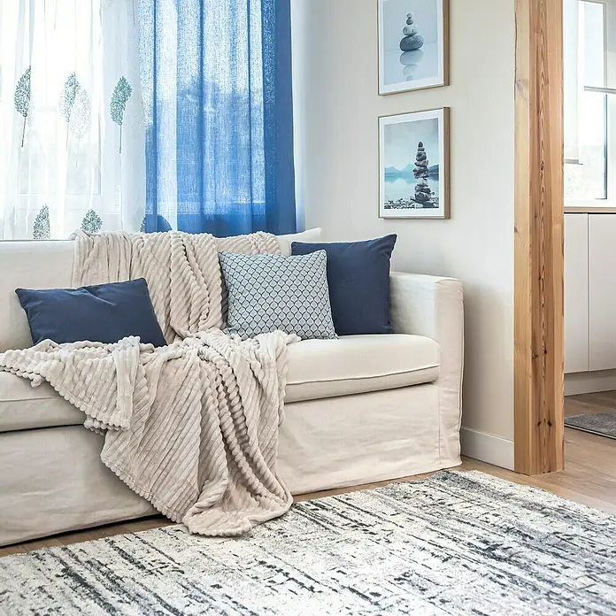 Manta para sofá Alpes - 180 x 200 cm, Poliéster, Beige - Primer plano de una sala de estar con un sofá color crema, cojines azules y una manta beige. Una alfombra estampada está sobre el piso de madera.