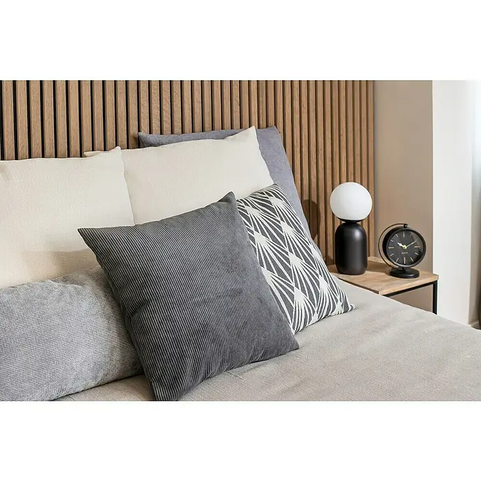 Cojín Pana - 45 x 45 cm, Poliéster, Gris, Cuadrado - Primer plano de una cama con almohadas elegantes en tonos grises y blancos, complementadas por una lámpara elegante y un reloj en una mesita de noche.