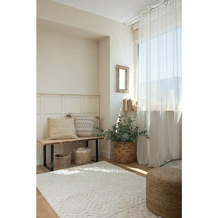 Cojín Dakira - 45 x 45 cm, Textil, Natural, Cuadrado - Acogedora habitación con una alfombra blanca, un banco de madera con cojines y un puf de mimbre, creando un ambiente cálido.