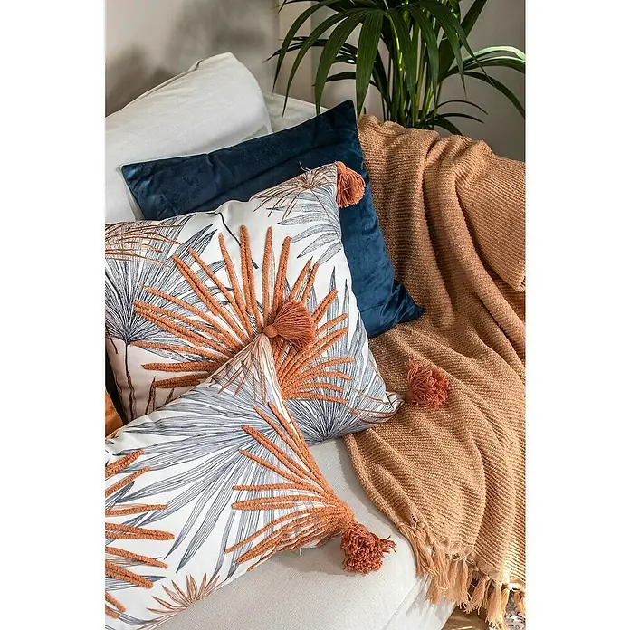 Manta para sofá Eira - 130 x 170 cm, Poliéster, Melocotón - Primer plano de cojines decorativos con diseño de hojas y un cojín azul marino en un sofá, junto a una manta marrón y una planta.