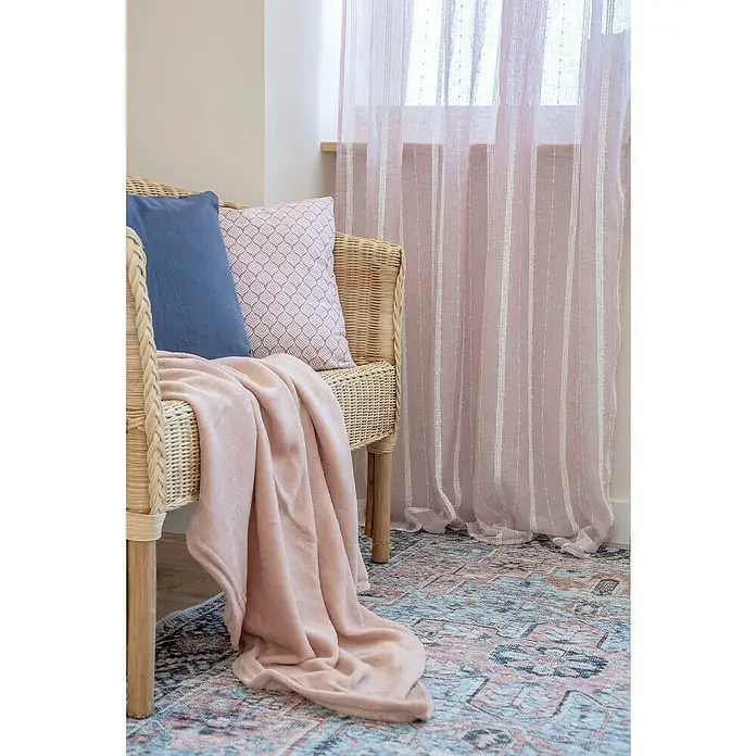 Manta para sofá Chicago - 130 x 150 cm, Poliéster, Rosa - Escena acogedora con una silla de ratán, cojines y una manta rosa suave, frente a una ventana con cortinas translúcidas.
