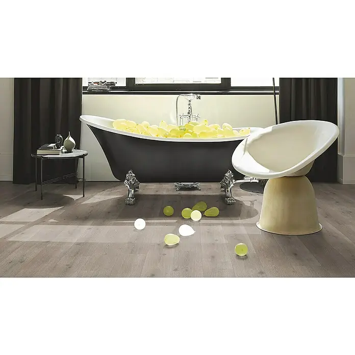 Parador Designbodenmuster Modular ONE Urban Grau - 200 x 194 x 8 mm, Landhausdiele - Ein elegantes Badezimmer mit freistehender Badewanne, gefüllt mit gelben Ballons, neben einem modernen Sessel.