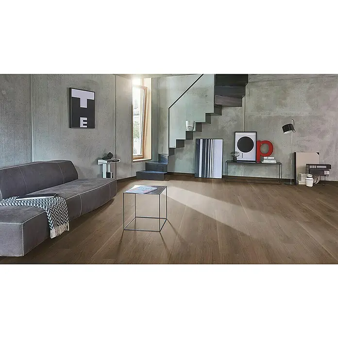 Parador Designboden Modular ONE Eiche Spirit Geräuchert1.285 x 194 x 8 mm, Landhausdiele, Eiche Spirit Geräuchert - Geräumiges Wohnzimmer mit braunem Holzboden, grauer Couch und Betonwänden, akzentuiert durch moderne Kunst und einem schlanken Beistelltisch.