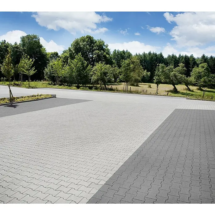 EHL Pflasterstein Doppel-TGrau, 16,5 x 10 x 8 cm, Randstein - Außenaufnahme eines gepflasterten Parkplatzes mit grauen Pflastersteinen, umgeben von Bäumen und unter blauem Himmel.