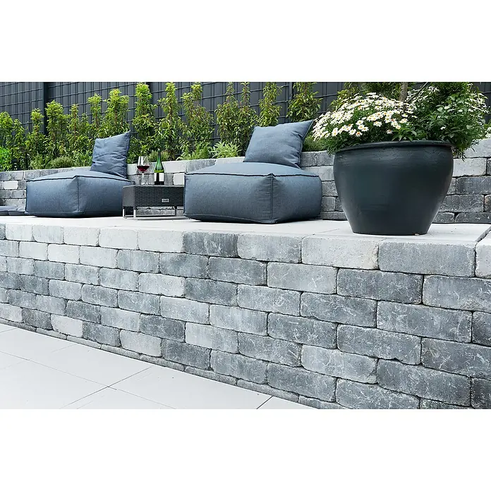 EHL Mauerstein Times Antik S - 20 x 20 x 7 cm, Grau/Anthrazit, Beton - Gemütliche Sitzecke im Freien mit grauen Sitzsäcken, Weinflasche und einem großen Blumentopf mit weißen Blumen auf einer Steinmauer.