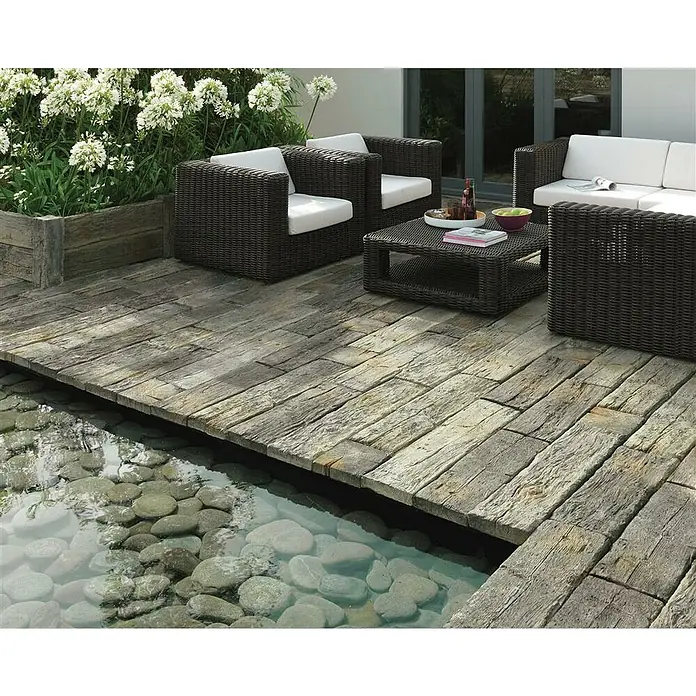 EHL Gehwegplatte Bahnschwelle22,5 x 22,5 x 4 cm, Naturbraun-grau, Beton - Rattanmöbel-Set auf Holzterrasse am Wasser. Beinhaltet Sessel, Sofa und Tisch, perfekt für stilvolle Entspannung im Freien.