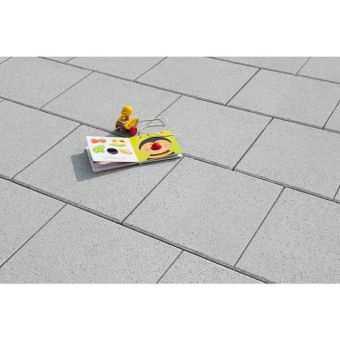 Diephaus Terrassenplatte Norman - 40 x 40 x 4 cm, Quarzgrau, Beton - Spielzeugente und Kinderbuch liegen auf dem grauen Pflaster. Das Buch zeigt eine lustige Biene und Früchte.