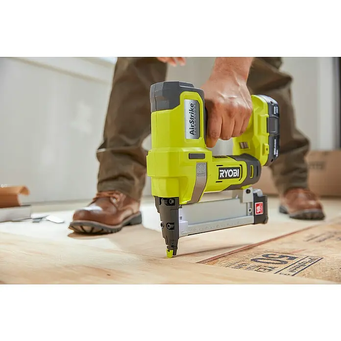 Ryobi ONE+ Akku-Tacker R18GS18-0 - 18 V, Li-Ionen, Ohne Akku - Nahaufnahme eines Ryobi AirStrike Akku-Nagelgeräts beim Eintreiben eines Nagels in ein Holzbrett, gehalten von einer Person mit Lederschuhen.