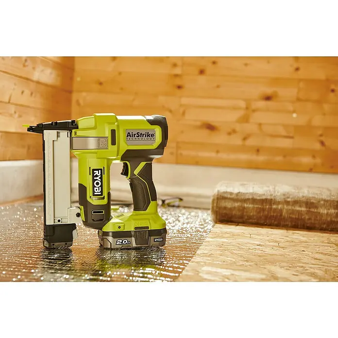 Ryobi ONE+ Maschinenpaket - Akku-Tacker R18GS18-0 + gratis Starter-Kit - Akkubetriebener Ryobi-Nagler mit AirStrike-Technologie auf einer reflektierenden Oberfläche mit Holzwand im Hintergrund.