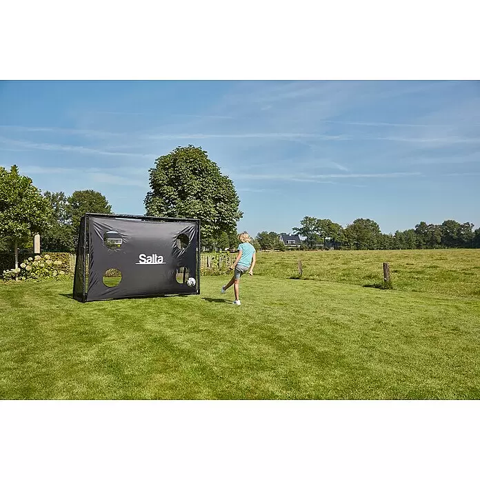 Salta Fußballtor - 300 x 90 x 200 cm, Schwarz - Ein Kind tritt einen Fußball auf ein schwarzes Salta Trainingsfußballtor mit Ziellöchern auf einem Rasenplatz.