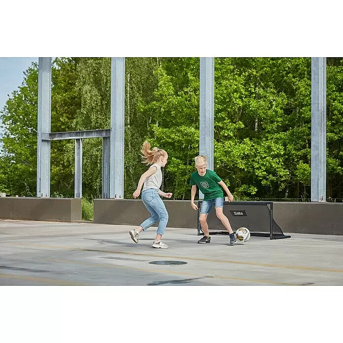 Salta Fußballtor Techniq - 150 x 60 x 60 cm, Schwarz - Zwei Kinder spielen Fußball mit einem schwarzen Salta tragbaren Minitor auf einem gepflasterten Platz.