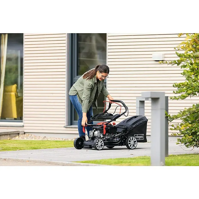 Scheppach Benzin-Rasenmäher PML46-150ES - 150 cm³, Schnittbreite: 46 cm, Hybrid-Start: Elektrostart & Seilzugstarter - Frau bereitet den Scheppach Benzin-Rasenmäher vor, um den Rasen zu mähen. Das Gerät ist kompakt und leicht zu bedienen.
