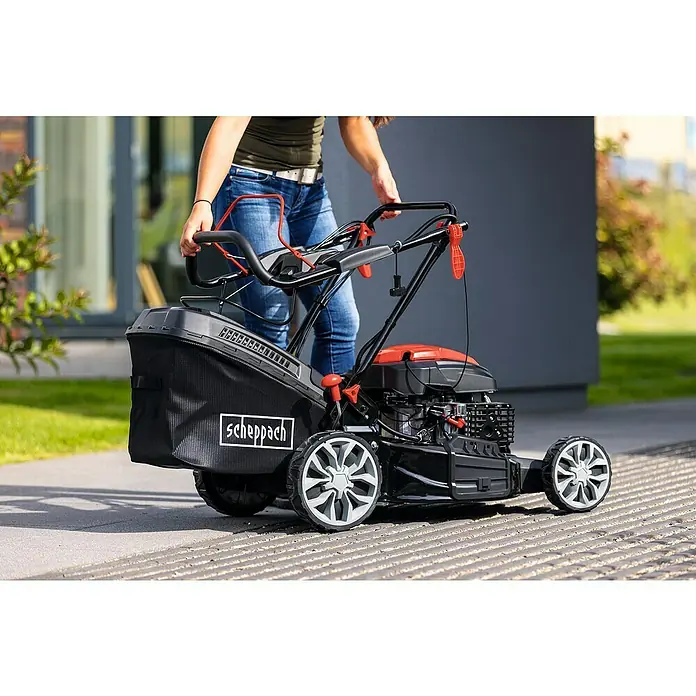Scheppach Benzin-Rasenmäher PML56-225ES - 224 cm³, Schnittbreite: 56 cm, Hybrid-Start: Elektrostart & Seilzugstarter - Eine Frau schiebt einen Scheppach Benzin-Rasenmäher mit Fangkorb auf einer gepflasterten Fläche. Das Gerät ist schwarz und orange.