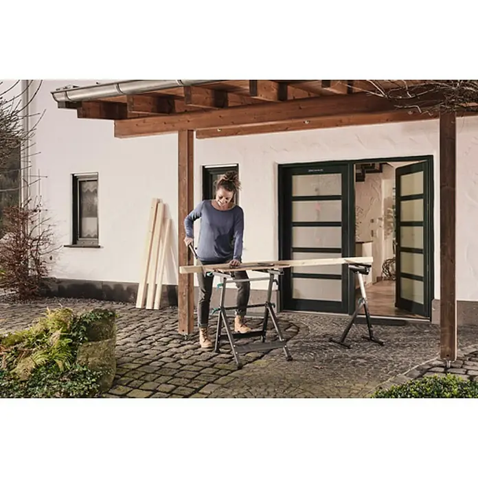 Wolfcraft Master Werkbank - L x B: 720 x 650 mm, Arbeitshöhe: 950 mm, Belastbarkeit: 120 kg - Eine Frau sägt ein Holzbrett auf einem Wolfcraft Werkzeugtisch im Freien, vor einem Haus mit Holzbalken und Pflastersteinen.