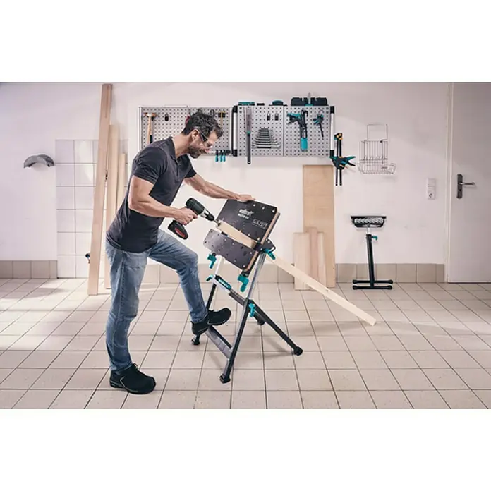 Wolfcraft Master Werkbank - L x B: 720 x 650 mm, Arbeitshöhe: 950 mm, Belastbarkeit: 120 kg - Ein Mann arbeitet mit einer Wolfcraft Master 600 Werkbank in einer Werkstatt, um Holz zu bearbeiten. Das Werkzeugbrett an der Wand hält verschiedene Werkzeuge bereit.