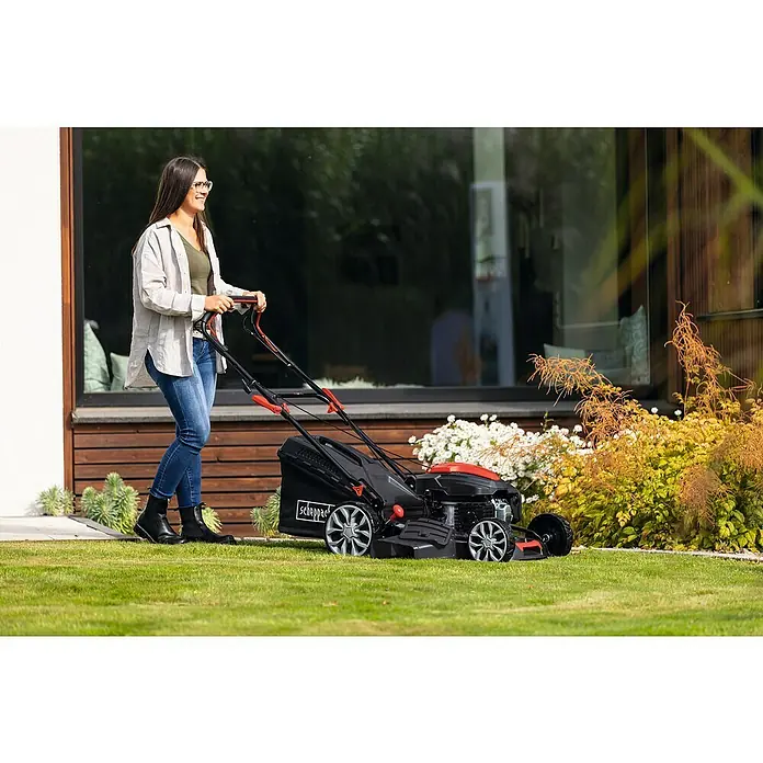 Scheppach Benzin-Rasenmäher PML53-225S - 224 cm³, Schnittbreite: 53 cm, Seilzugstarter - Eine Frau benutzt einen Scheppach Rasenmäher im Garten, um das Gras zu schneiden. Der Rasenmäher ist schwarz-rot und wirkt leistungsstark.