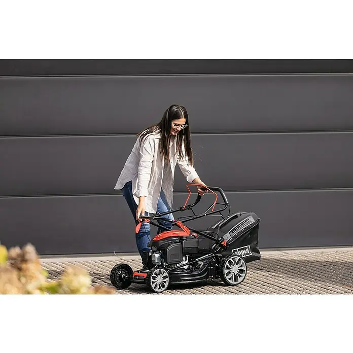 Scheppach Benzin-Rasenmäher PML53-225S - 224 cm³, Schnittbreite: 53 cm, Seilzugstarter - Eine Frau steht vor einem Garagentor und schiebt einen Scheppach Benzin-Rasenmäher mit rotem Griff.