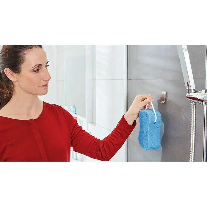 Tesa Powerstrips Wandhaken Zoom Small - 2 Stk., Rechteckig, Metall - Eine Frau hängt einen blauen Badeschwamm mit einer weißen Schnur an einen silbernen Haken im Badezimmer.