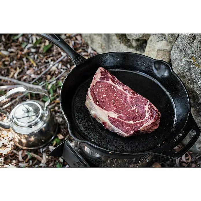 Petromax Grillpfanne - Ø x H: 30 x 4,5 cm, Gusseisen - Ein rohes Ribeye-Steak, gewürzt mit Salz, liegt in einer schwarzen gusseisernen Pfanne, bereit zum Kochen im Freien auf einem Campingkocher.