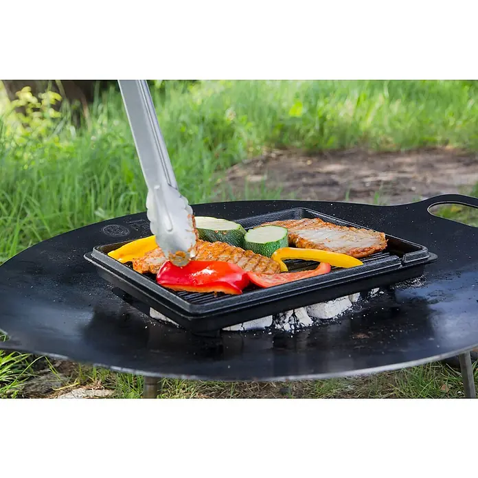 Petromax Auflaufform - L x B x H: 34 x 24 x 14,5 cm, Gusseisen, 4,5 l - Gemüse und Fleisch werden auf einer Grillplatte zubereitet. Eine Grillzange wird benutzt, um das Essen zu wenden, während es brutzelt.