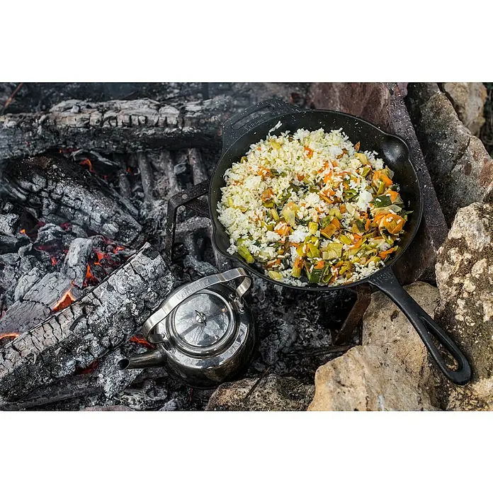 Petromax Grillpfanne - Ø x H: 30 x 4,5 cm, Gusseisen - Eine gusseiserne Pfanne mit Reisgericht steht über einem Lagerfeuer, daneben ein Wasserkessel. Ideal zum Kochen im Freien.