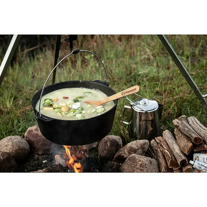 Petromax Dutch Oven - Ø x H: 42 x 20 cm, Gusseisen, 10,8 l, Planer Boden - Ein Petromax Dutch Oven hängt über einem Lagerfeuer, gefüllt mit einer herzhaften Suppe und einem Holzlöffel.