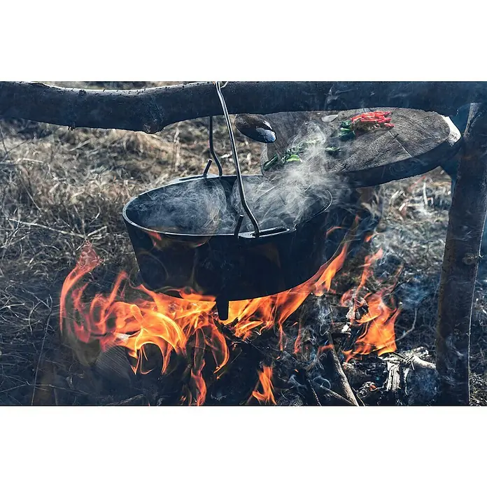 Petromax Dutch Oven - Ø x H: 36,4 x 20 cm, Gusseisen, 5,5 l, Mit Füßen - Über einem offenen Feuer hängt ein dampfender Kochtopf, der rustikales Outdoor-Kochen und die Einfachheit der Zubereitung von Speisen im Freien zeigt.