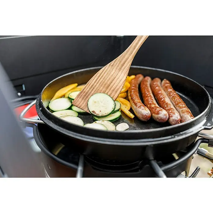Petromax Grillgutwender - Länge: 32 cm, Kirschholz - Eine gusseiserne Pfanne mit Würstchen, Zucchini und Paprika wird mit einem Holzspatel gewendet.