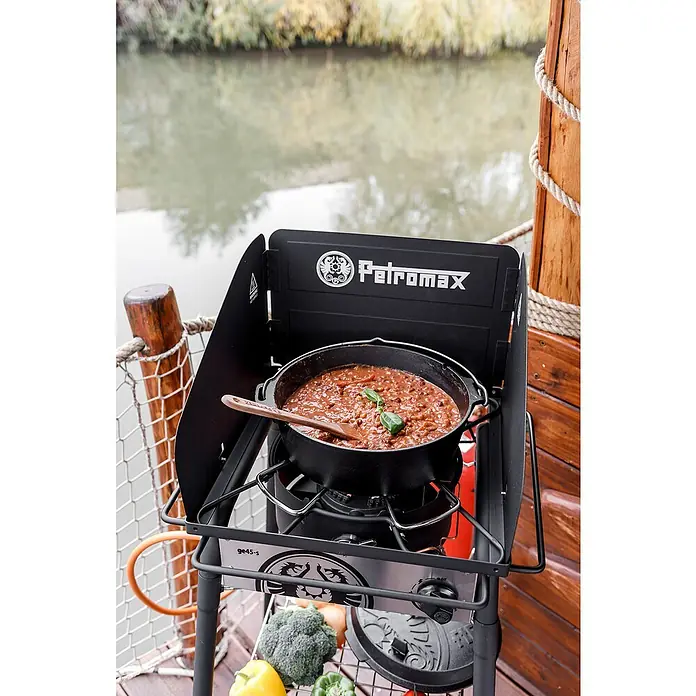 Petromax Dutch Oven - Ø x H: 36,4 x 23,2 cm, Gusseisen, 7,5 l, Mit Füßen - Ein Petromax Kocher mit Windschutz und einem Eintopf, der auf einem Schiff zubereitet wird. Gemüse befindet sich unter dem Kocher.