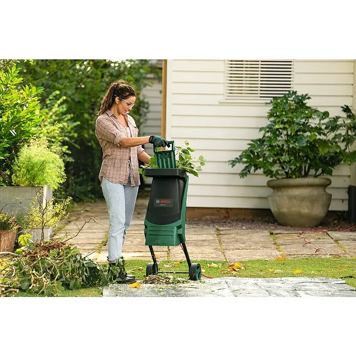 Bosch Elektro-Häcksler AXT RAPID 2200 - Max. Aststärke: 4 cm, 2.200 W - Eine Frau benutzt einen Bosch Gartenhäcksler, um Gartenabfälle zu zerkleinern. Das Gerät steht auf einer Plane im Garten.