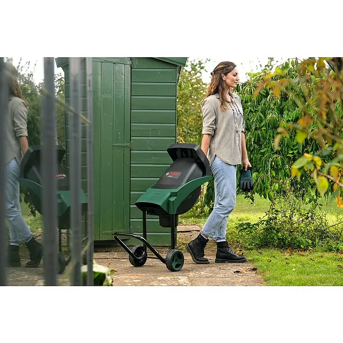 Bosch Elektro-Häcksler AXT RAPID 2200 - Max. Aststärke: 4 cm, 2.200 W - Eine Frau steht neben einem Bosch Gartenhäcksler auf Rädern, ideal für die Gartenarbeit und zum Zerkleinern von Ästen.