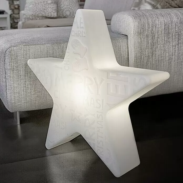8 Seasons Design Shining LED-Stern Star Merry Christmas - Weiß, Durchmesser: 60 cm, Polyethylen, 9 W, Lampenfassung: E27 - Große weiße transluzente Stern-LED-Weihnachtsdekoration mit "MERRY CHRISTMAS!" Schriftzug, leuchtend auf dem Boden vor einem Sofa.