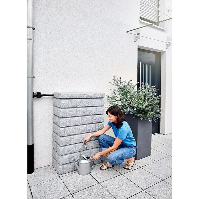 Garantia Exklusive Line Regenspeicher Rocky Junior - 300 l, Light Granit - Frau befüllt eine graue Gießkanne mit einem grauen Regentonne mit Hahn, der an einer weißen Hauswand steht.
